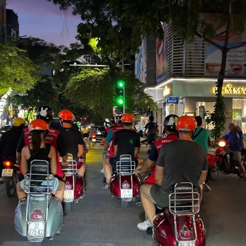 En gruppe mennesker med hjelme kører på scootere om natten i en by og venter ved et grønt trafiklys.