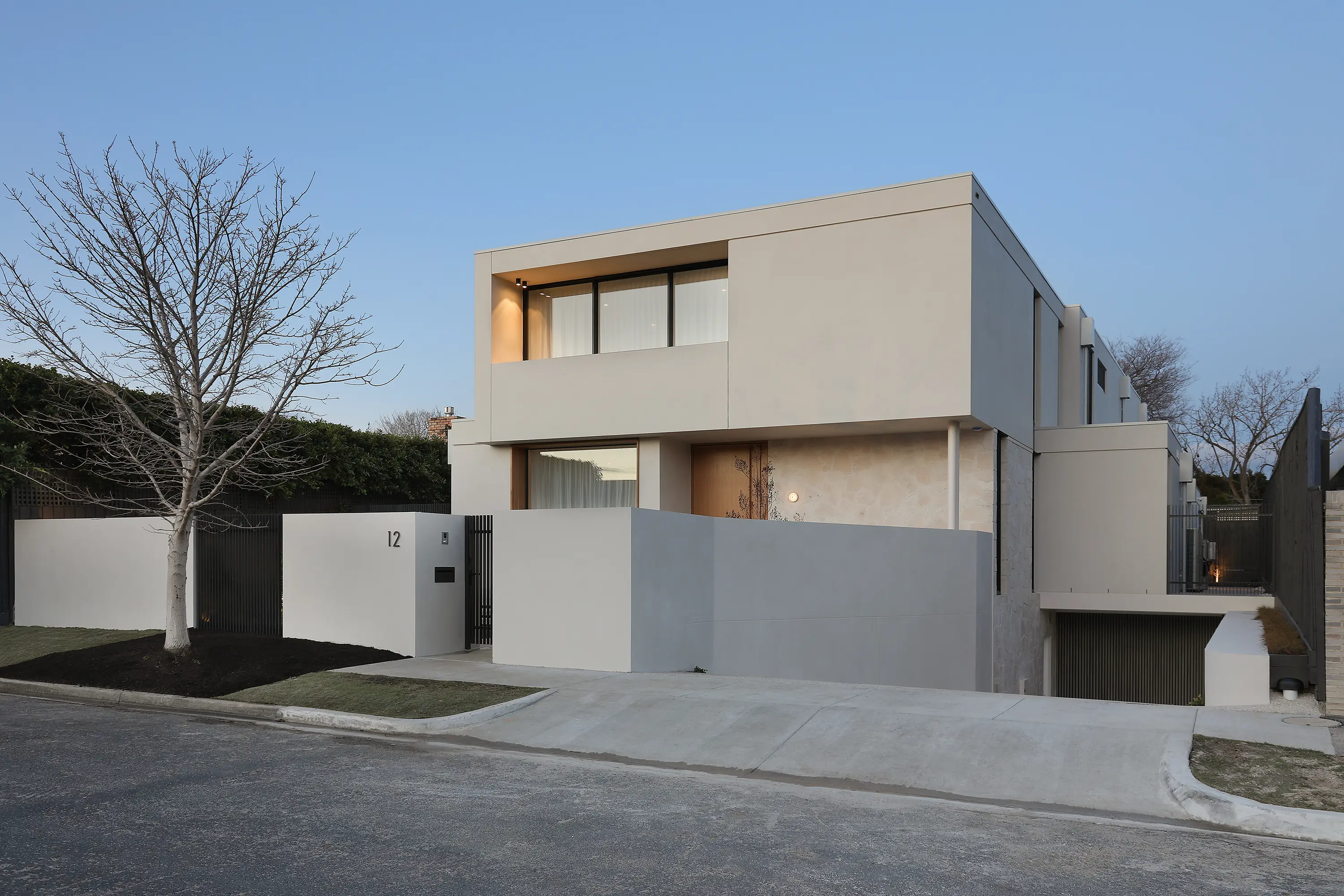 Front facade of Brighton Residence at dusk, a two-storey grey-rendered minimalist home with a timber entry door, rendered boundary wall and a bare tree on the nature strip