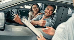 A man in the driver's seat of a car gives a thumbs up while a woman smiles beside him, as a person in the foreground holds a clipboard.