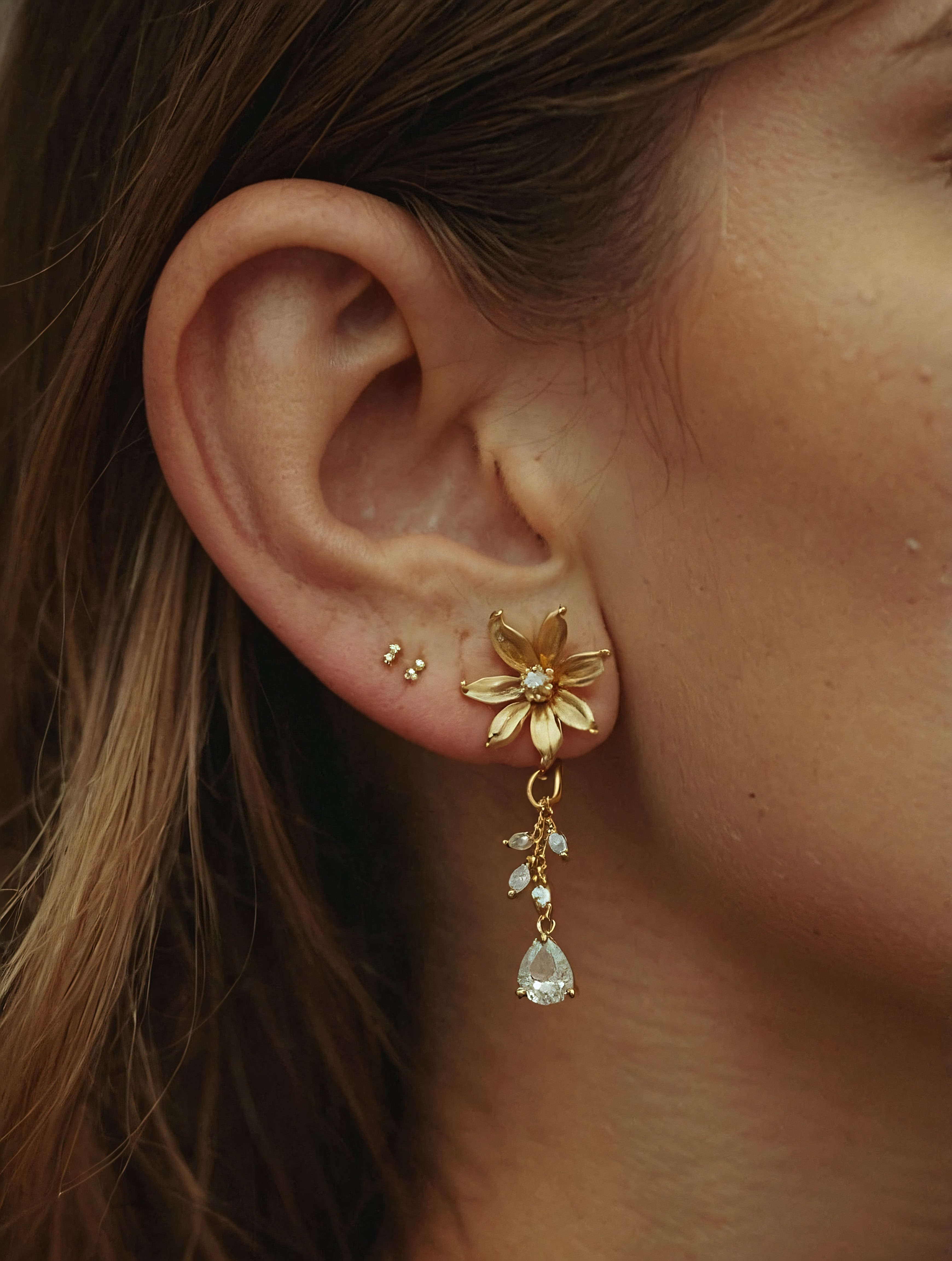 A close-up of a woman’s ear adorned with elegant, gold floral earrings, with a gemstone teardrop hanging below.