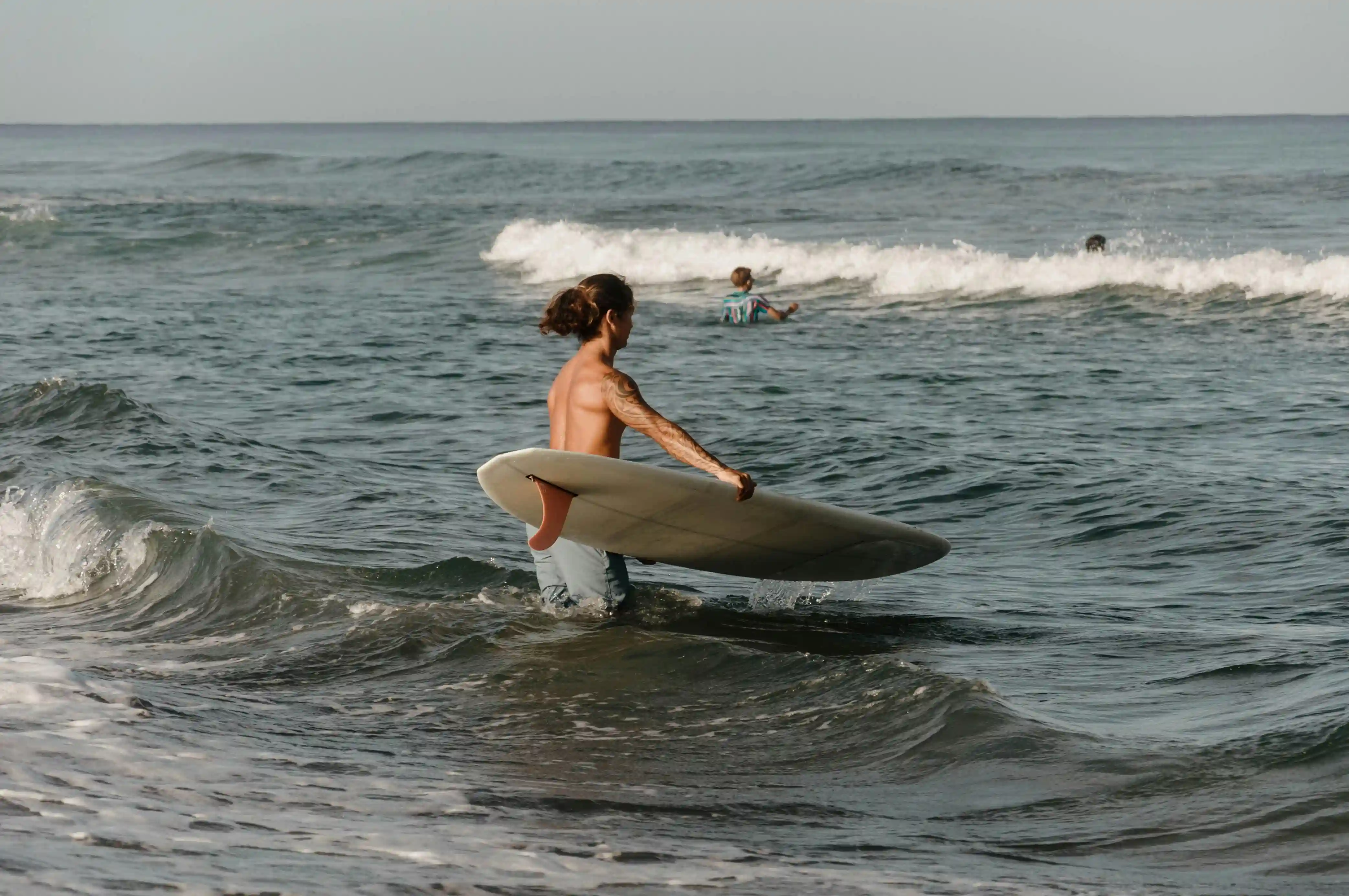 Surfer walking into the ocean carrying a surfboard as small waves break along the shore.