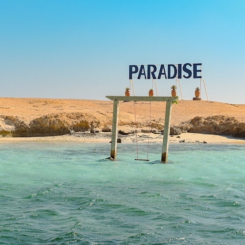 Wooden swing set in shallow turquoise water near a sandy shore, with a "PARADISE" sign and potted plants on top.