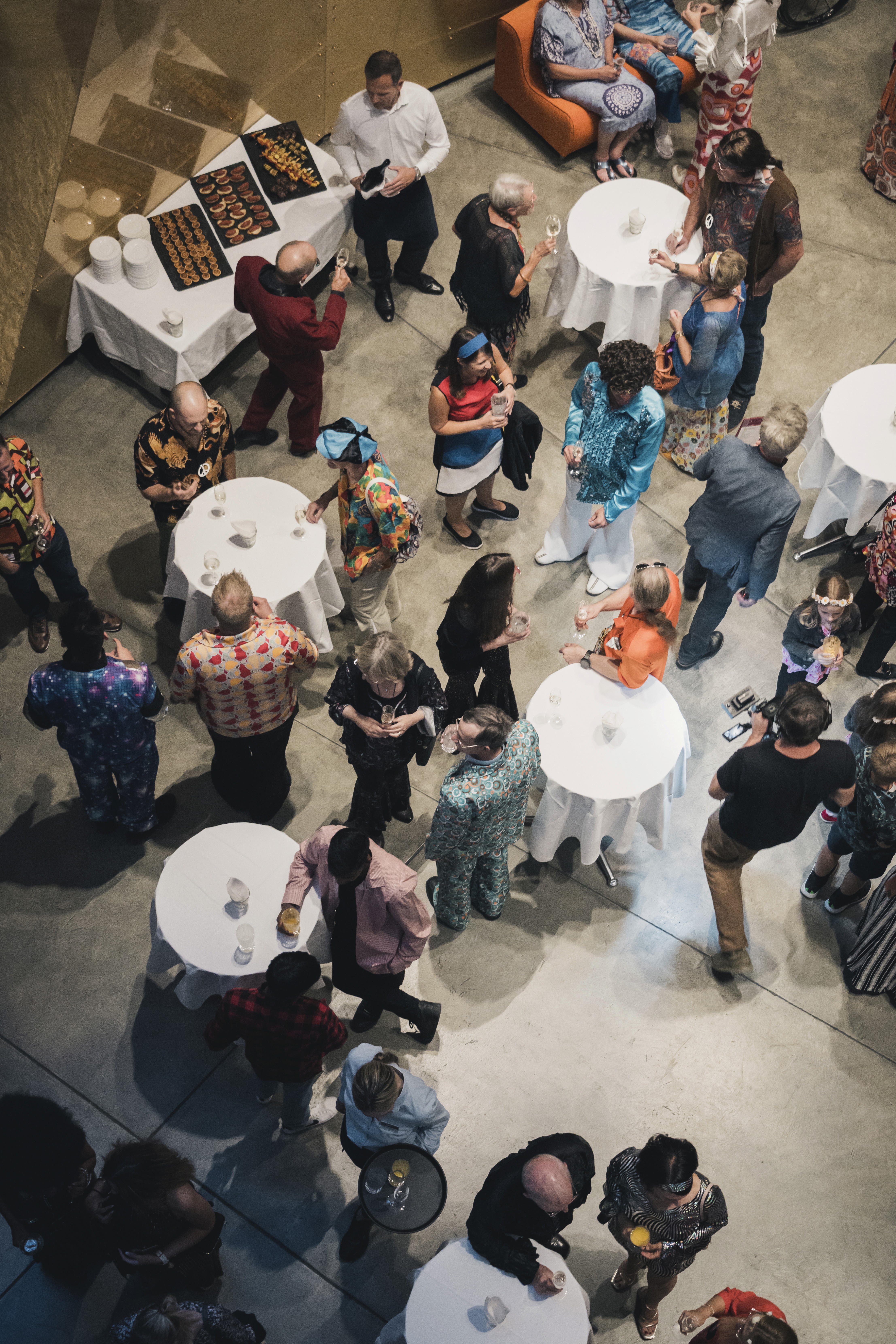 Das Foto zeigt aus der Vogelperspektive eine festliche Abendveranstaltung in einem großen Raum mit glattem Beton- oder Steinboden. Zahlreiche Gäste stehen in lockeren Gruppen an weiß gedeckten, runden Stehtischen zusammen, unterhalten sich und halten Gläser in den Händen. Auffällig ist, dass viele der Anwesenden farbenfrohe Kostüme oder Outfits im Retro- bzw. 70er-Jahre-Stil tragen, darunter schimmernde Paillettenoberteile, Schlaghosen, Stirnbänder und bunte Muster. Oben links im Bild befindet sich ein rechteckiger Tisch mit einem kleinen Buffet aus Häppchen, hinter dem Servicepersonal in weißen Hemden und schwarzen Hosen steht. Im unteren Bildbereich sieht man zudem eine Servicekraft, die ein Tablett mit Getränken trägt.