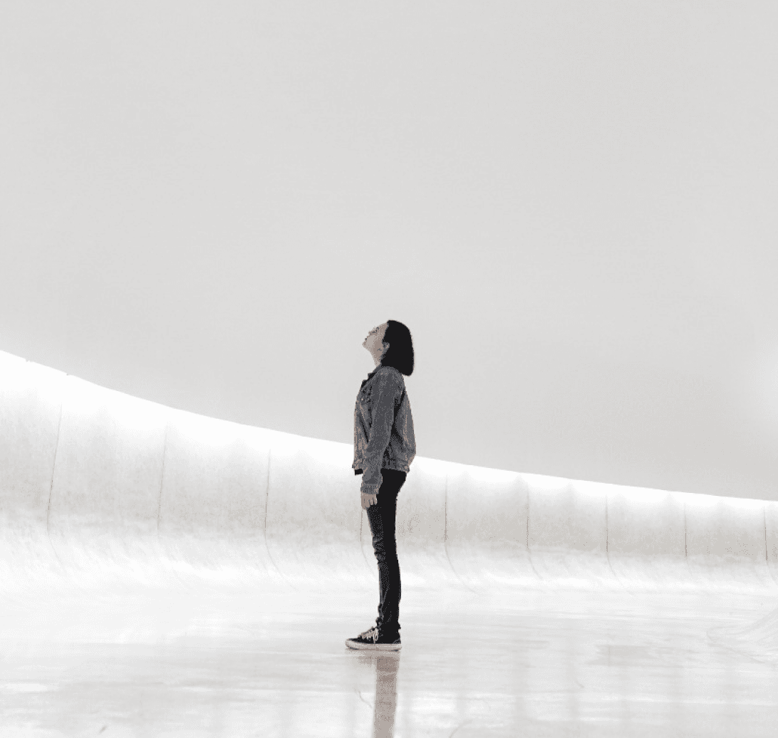 Girl standing in a bright, minimalist white room, gazing upward as if lost in thought.