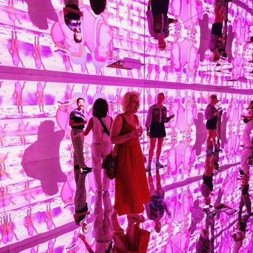 People standing in a room with reflective floors and walls, surrounded by vibrant pink and purple light projections.