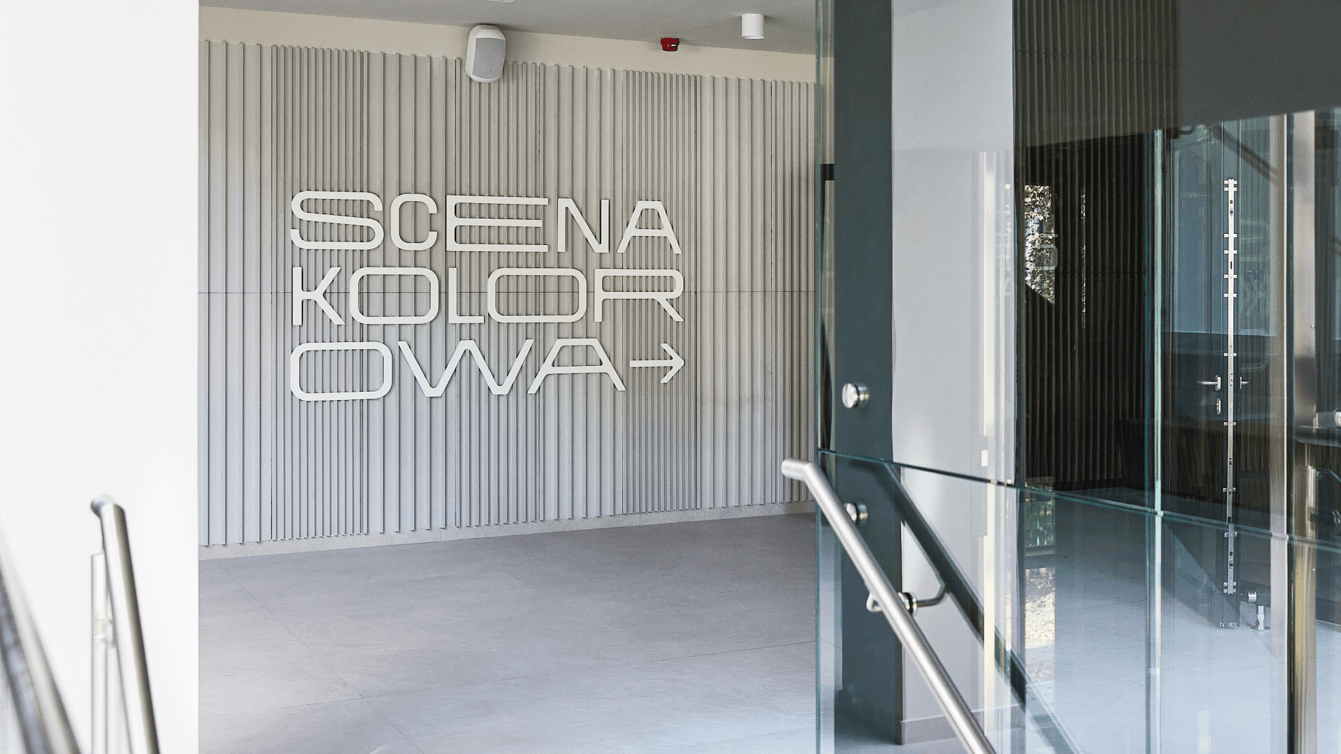 Biały napis „SCENA KOLOROWA” ze strzałką na jasnej ścianie, widziany pod kątem z korytarza.