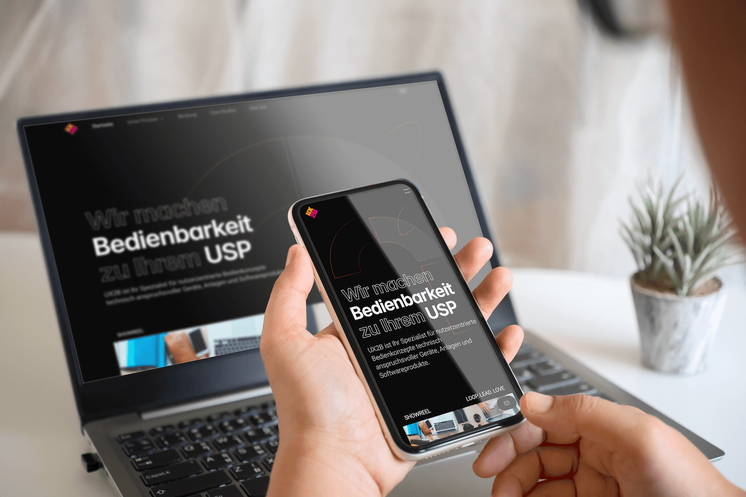 Hand hält Smartphone mit Website-Startseite vor aufgeklapptem Laptop mit derselben Website im Hintergrund