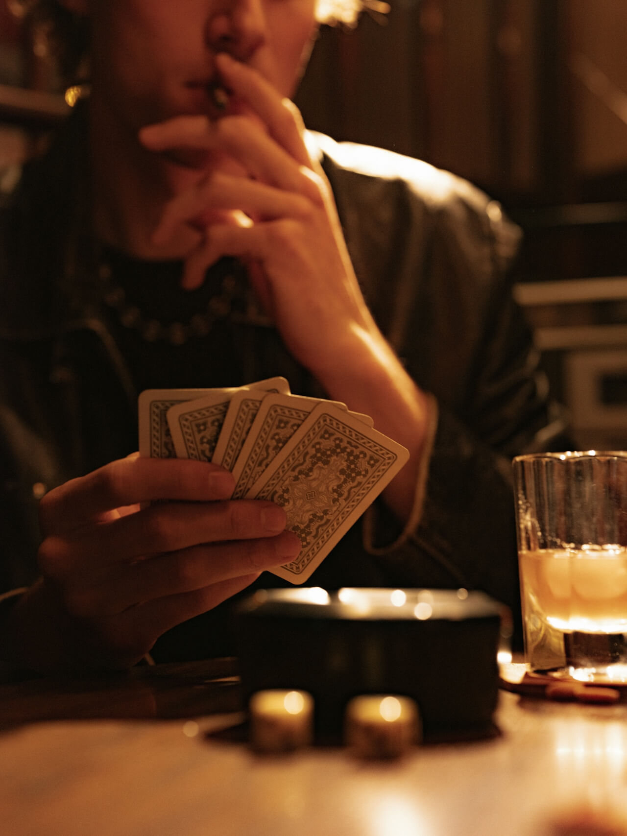 A person holding playing cards with a contemplative expression, smoking a cigarette. A glass and ashtray are on the dimly lit table, creating a tense mood.