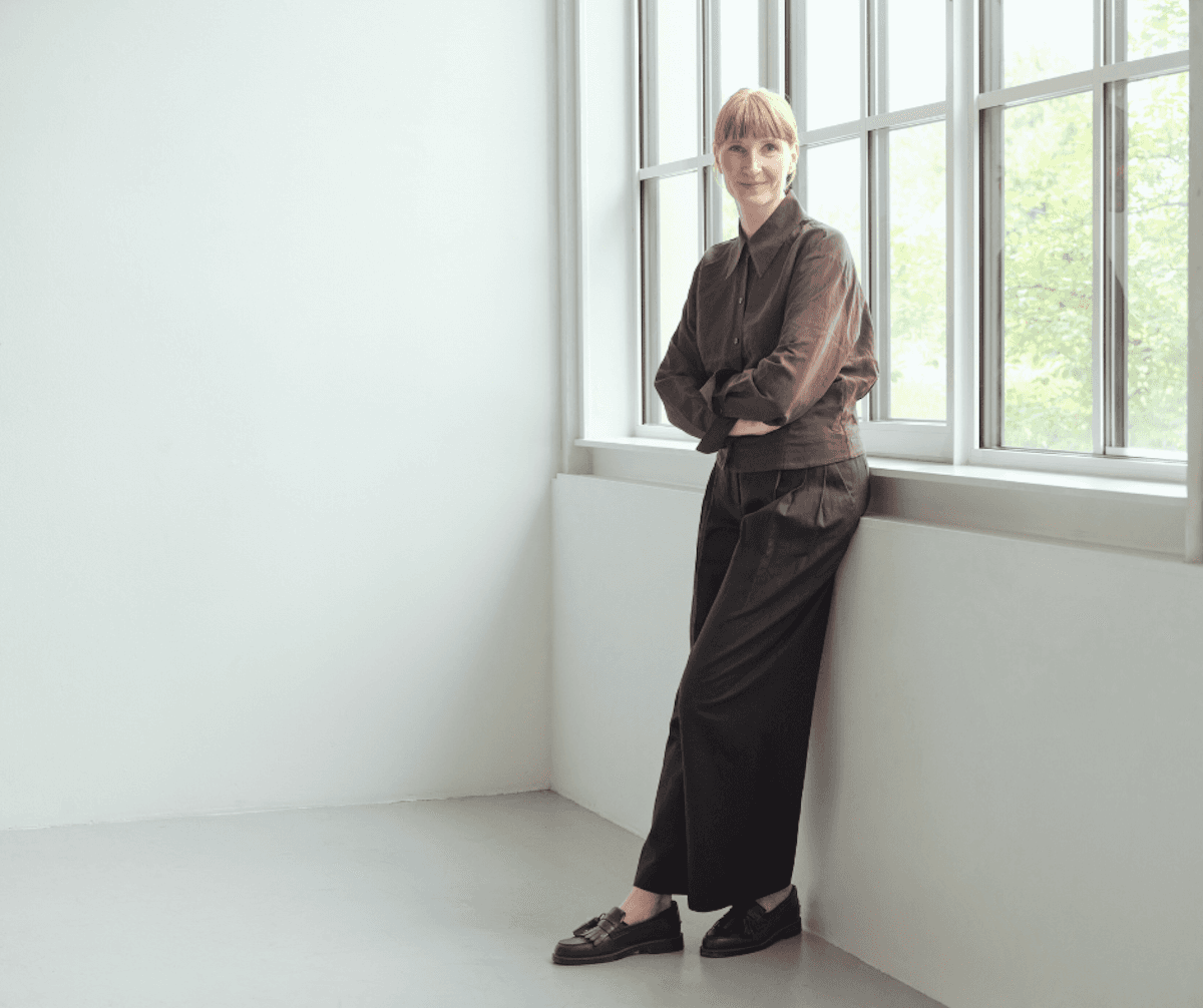 A person with short blonde hair, wearing a brown outfit, leans against a windowsill in a bright room. They appear relaxed and content.