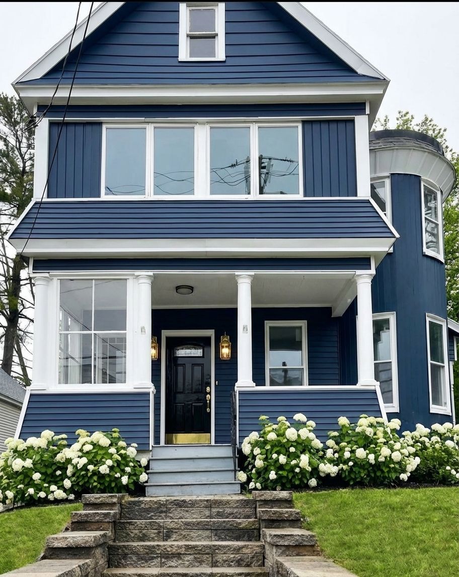 Deep navy siding with bright white trim, glossy black front door, brass accents, and white hydrangea foundation plantings