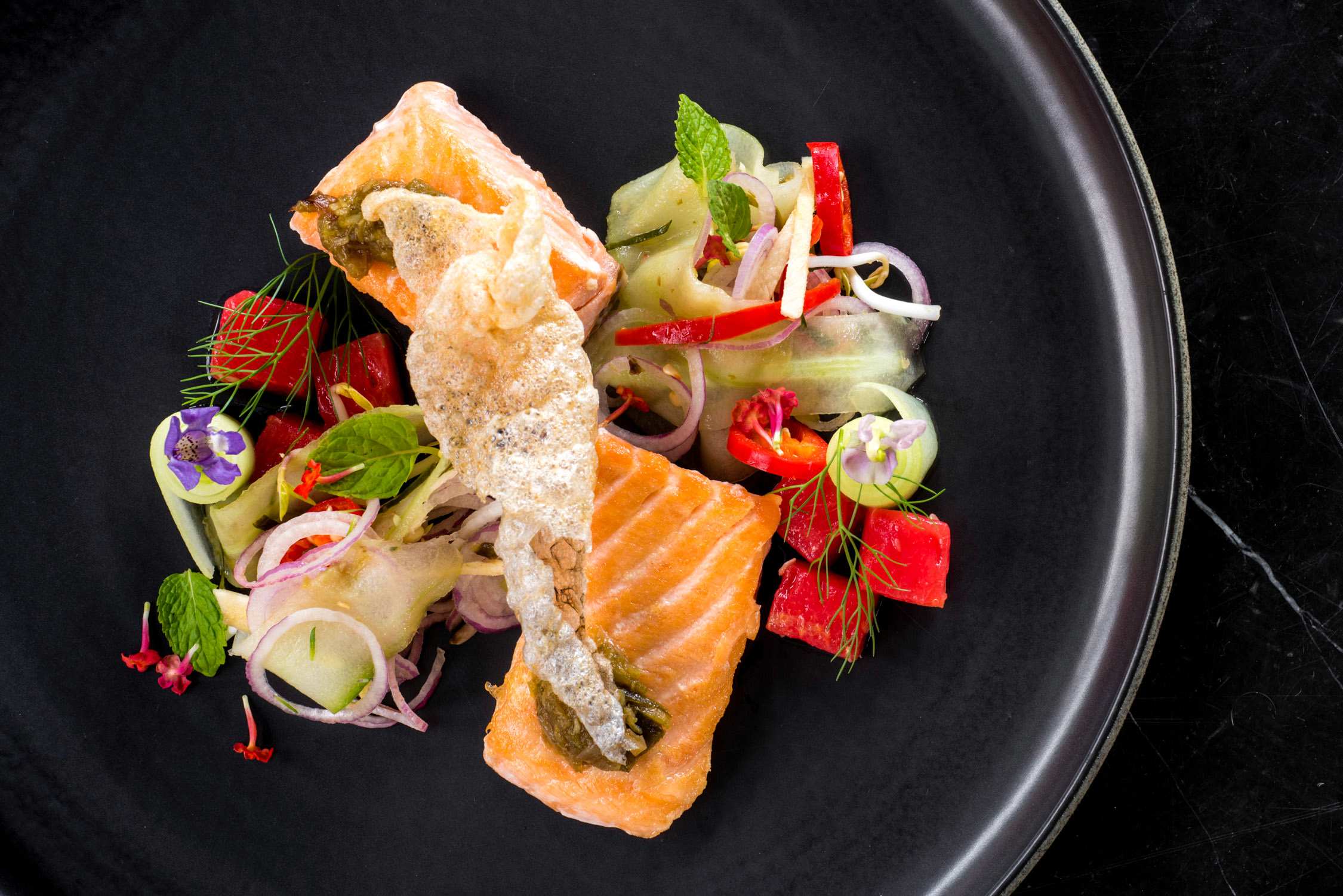 A piece of seared salmon topped with a crispy tuile sits on a black plate garnished with cucumber, radish, and herbs.