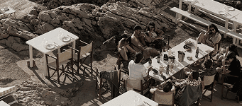 A relaxed seaside lunch with friends gathered around tables on rocky terrain, radiating warmth, joy, and effortless Mediterranean charm.  