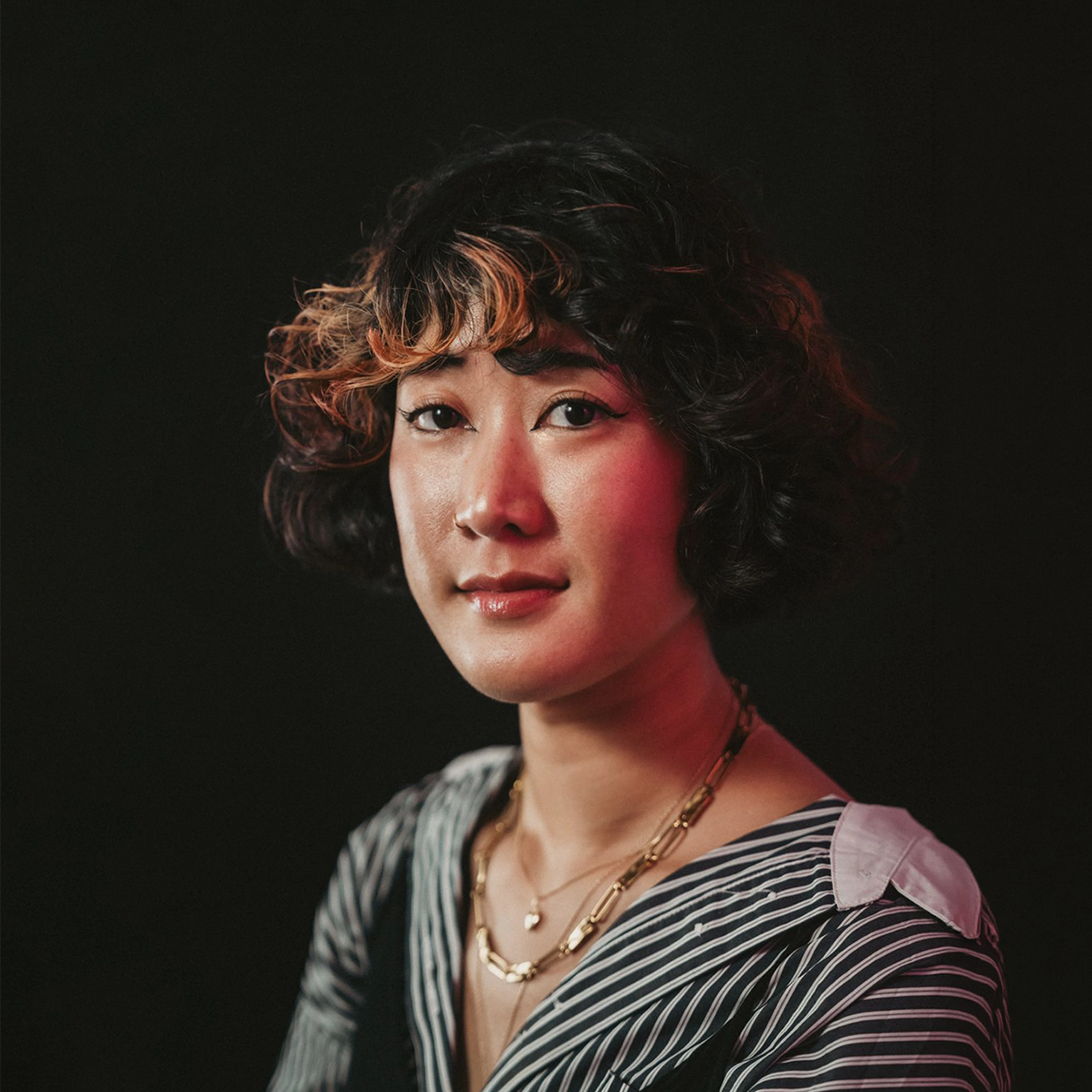 Photo portrait de profil de la designer et directrice artistique Diana SOM. Une jeune femme aux cheveux noirs bouclés courts avec des reflets cuivrés, portant une chemise rayée noire et blanche et des colliers dorés. Le portrait est réalisé sur un fond noir avec un éclairage studio doux et chaleureux.