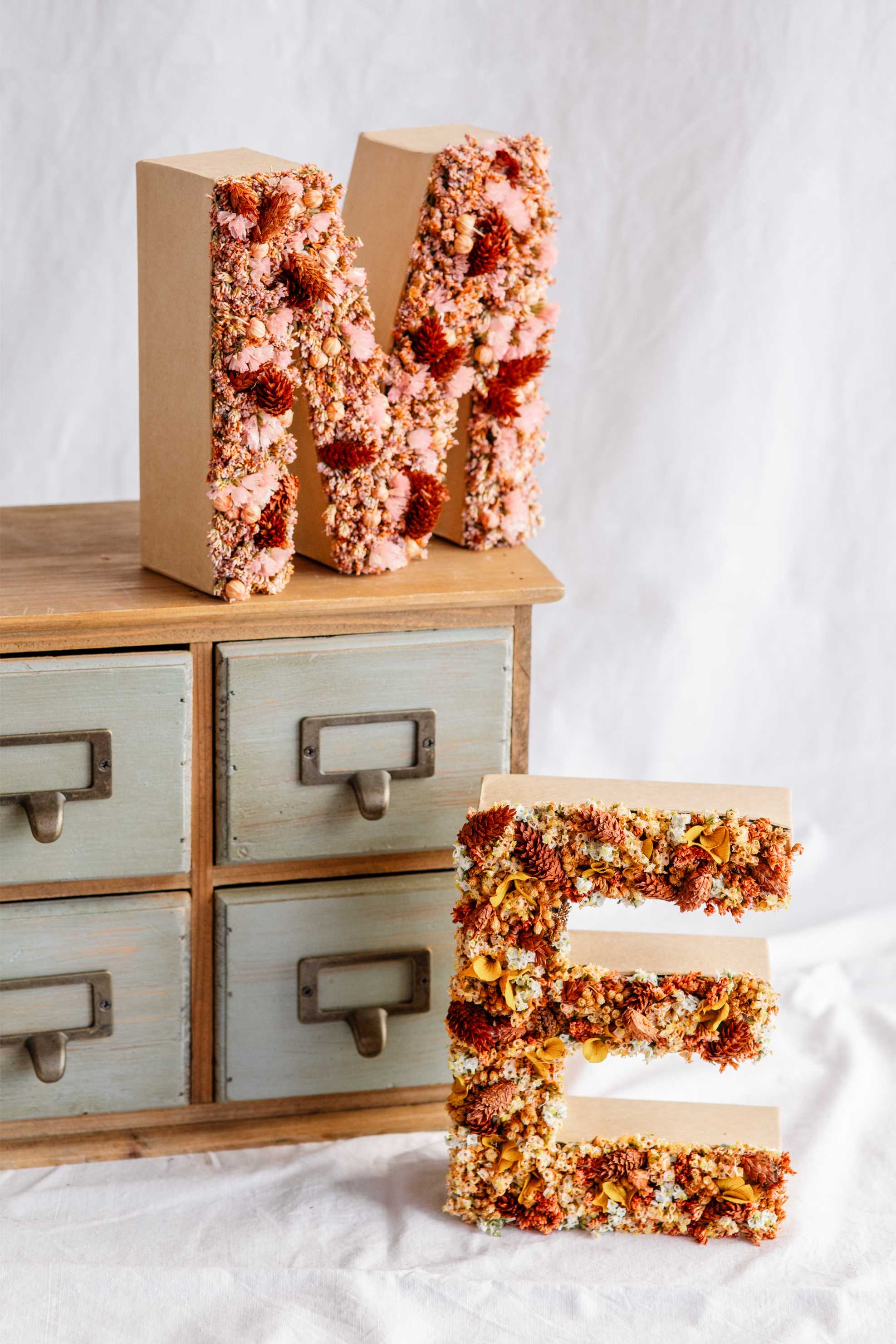 Lletres decoratives M i E cobertes amb flors seques de tons suaus i càlids, sobre una calaixera de fusta vintage.