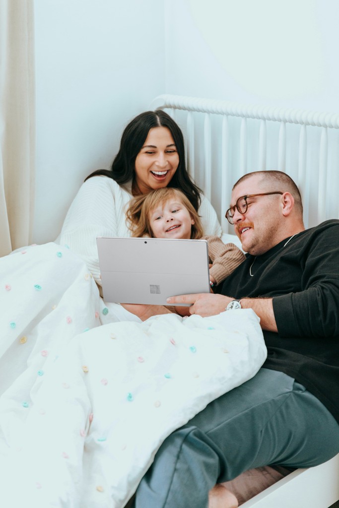 Father, mother, and daughter lying in bed, laughing and enjoying time together while watching their deceased loved one through Glimpse on a laptop.