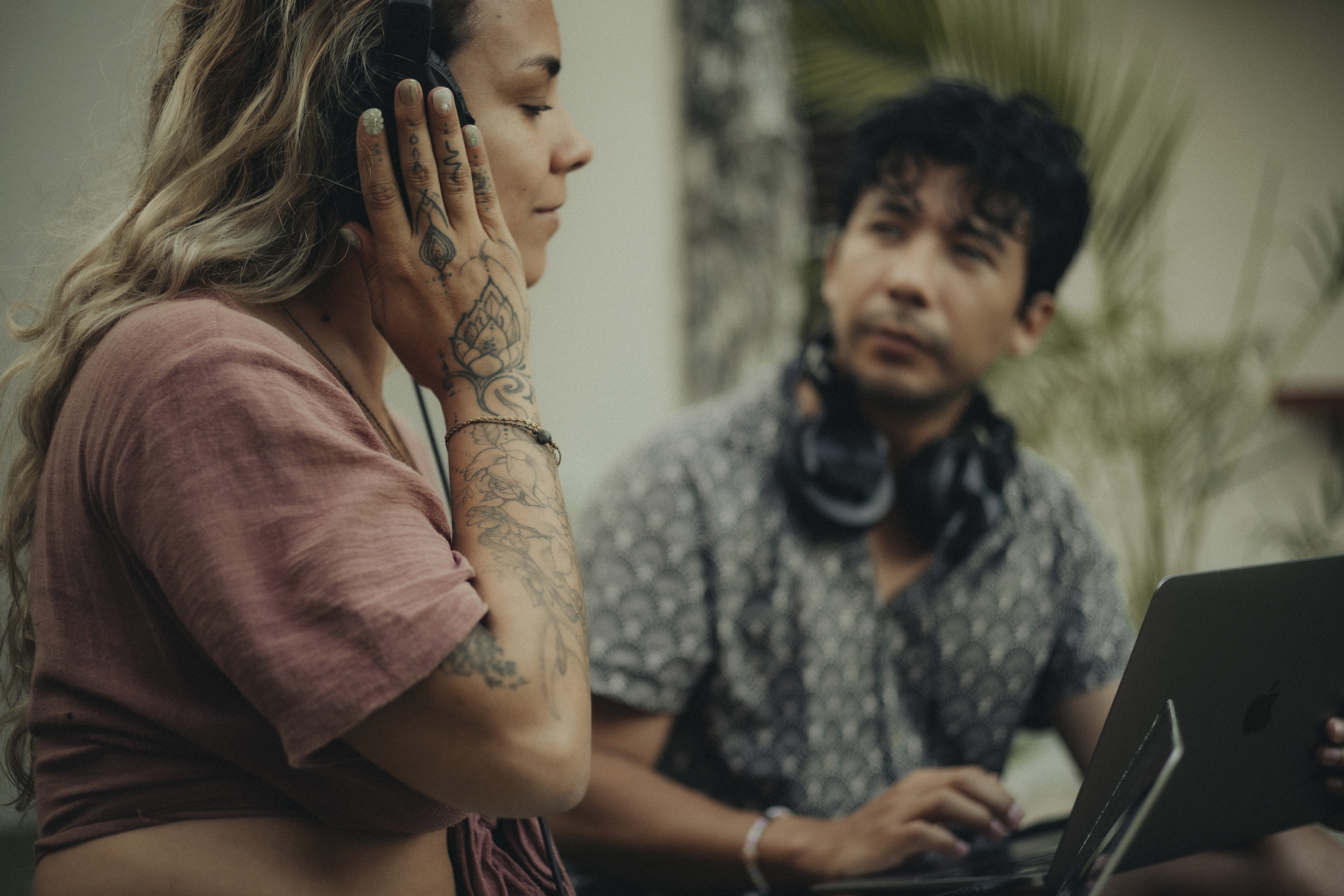 Girl wearing headphones with one hand on her ear, while a slightly blurred guy in the background looks at her and holds a laptop on his knees.