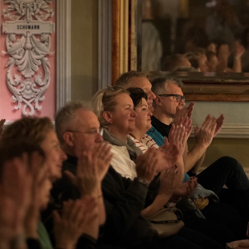 Audience members seated and applauding in an ornately decorated room with a sign labeled "Schumann" on the wall.