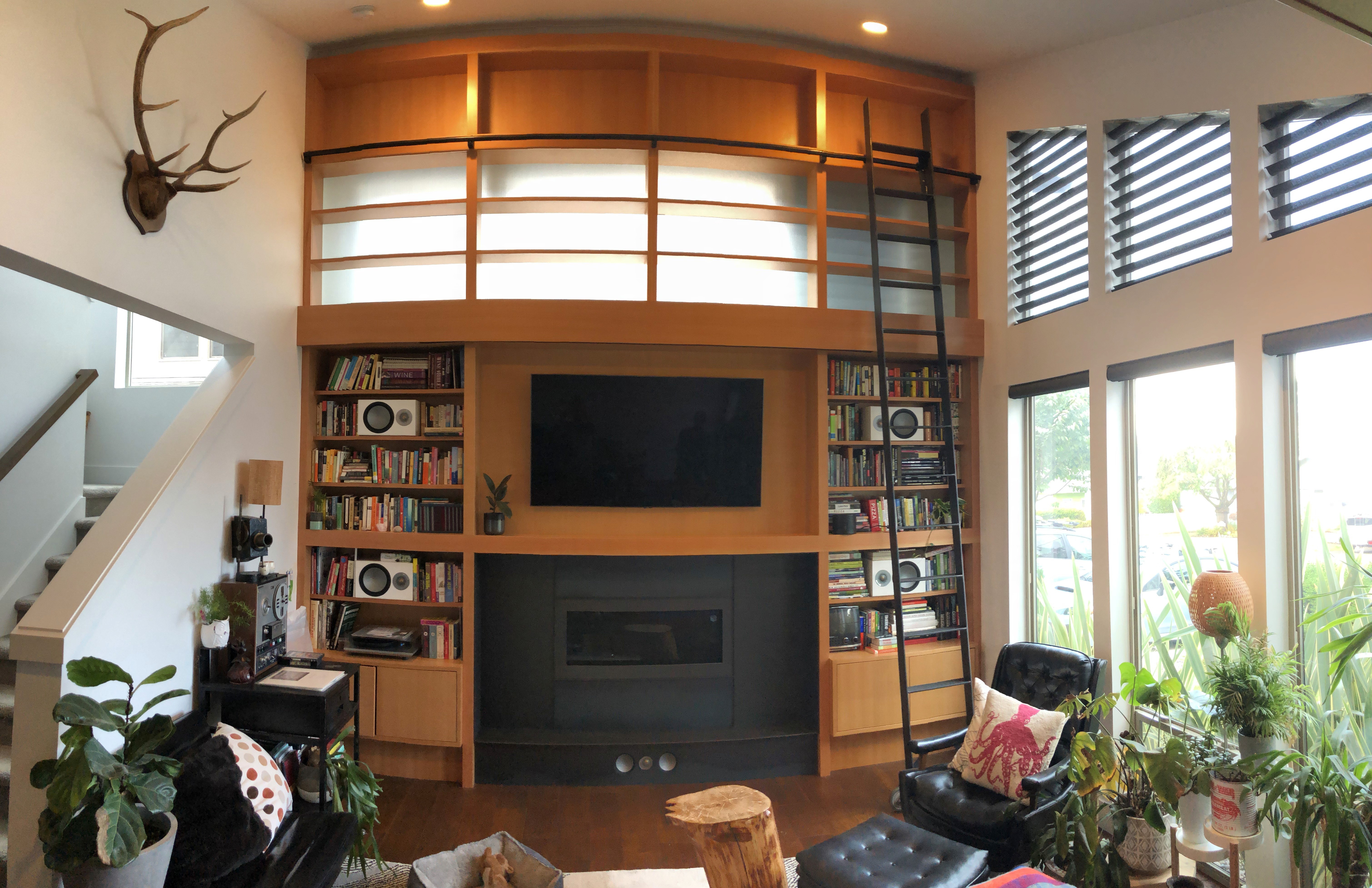 A floor-to-ceiling wooden bookcase and a rolling ladder with inset TV and fireplace sit below frosted windows in a den.