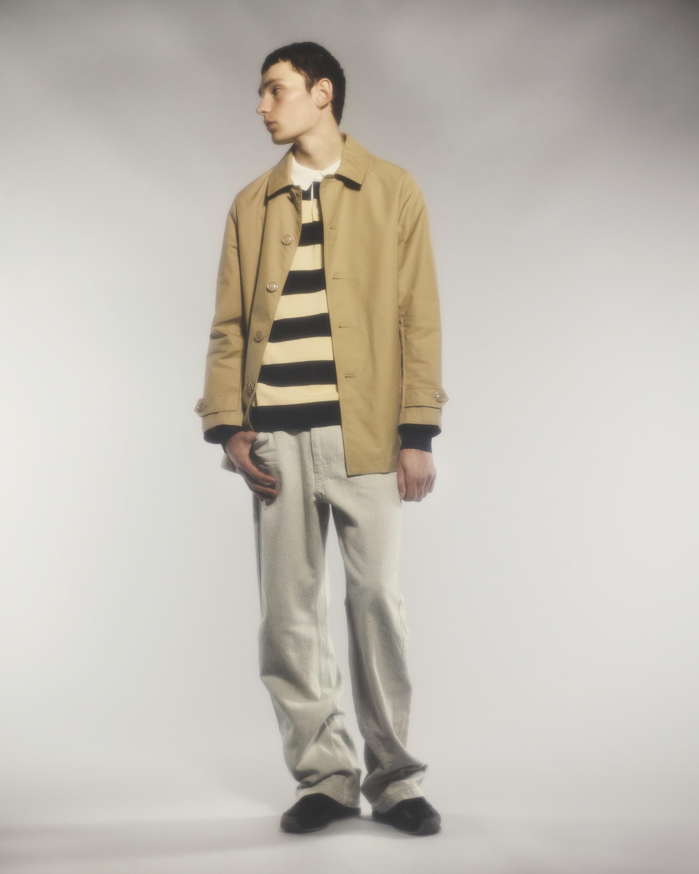 Sur un fond neutre,un mannequin porte un imperméable beige, un polo rayé et un pantalon large