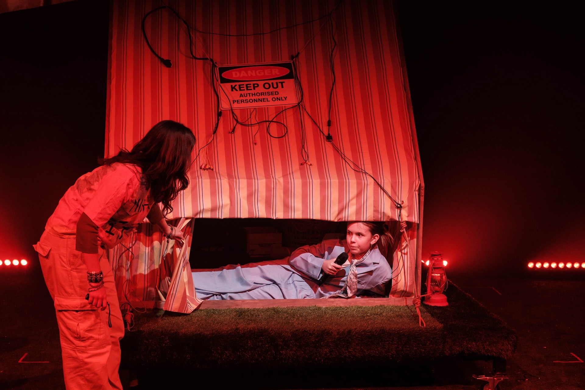A performer in a blue jumpsuit lies inside a striped makeshift tent, holding a microphone and peering out. Another performer lifts the edge of the tent flap to look in. A “Danger: Keep Out – Authorised Personnel Only” sign and hanging wires decorate the tent, all lit in dramatic red lighting.