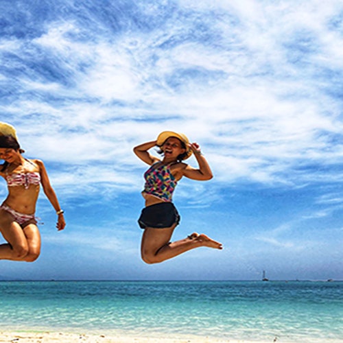 两名身穿泳衣和帽子的女性在海滩上跳跃，背景是清澈的蓝色海水和局部多云的天空。