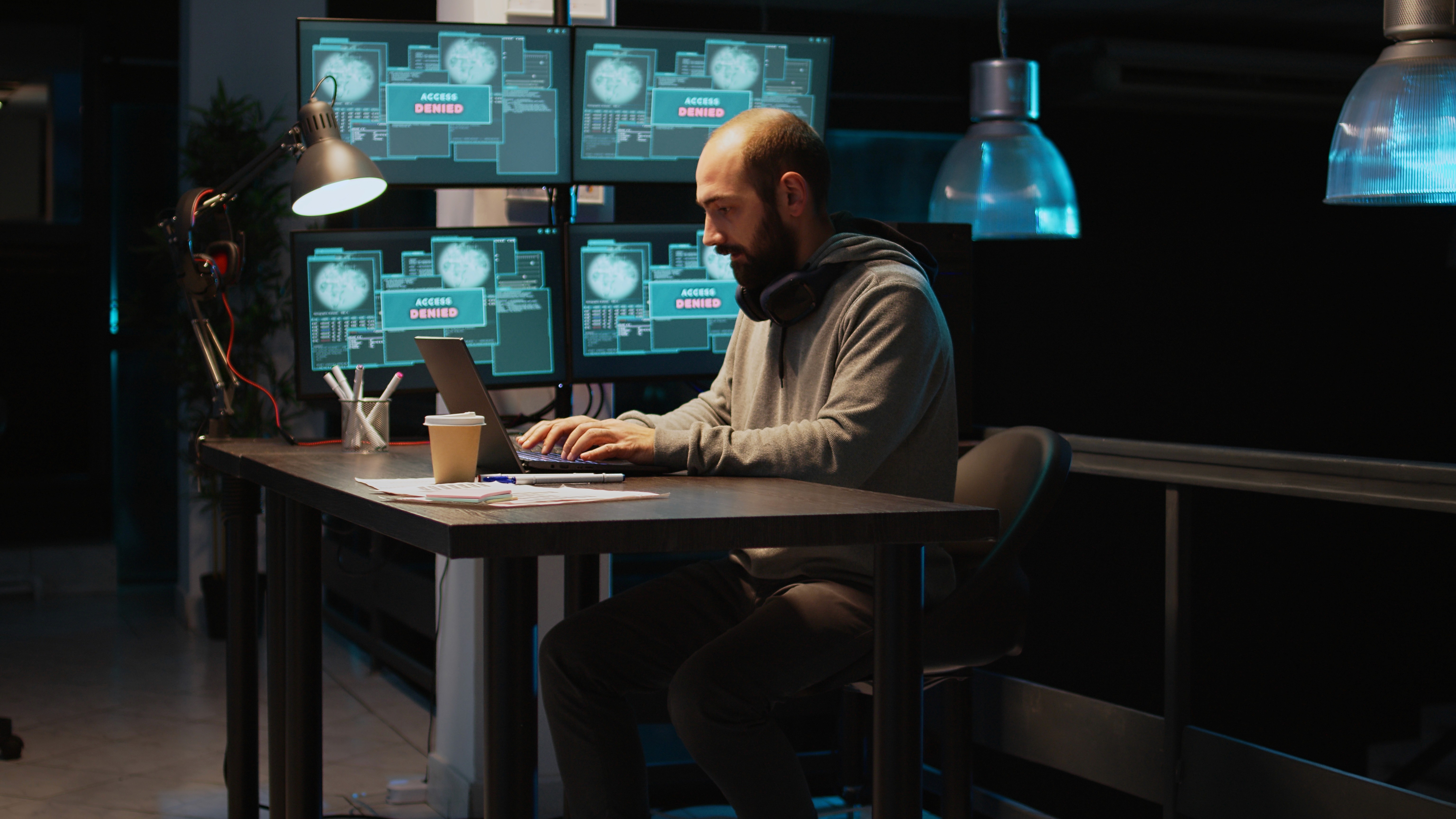 Ein Mann sitzt nachts in einem dunklen Büro vor einem Laptop und arbeitet konzentriert. Hinter ihm zeigen vier Monitore die rote Warnmeldung "ACCESS DENIED" (Zugriff verweigert) und symbolisieren eine kritische IT-Sicherheitskrise.