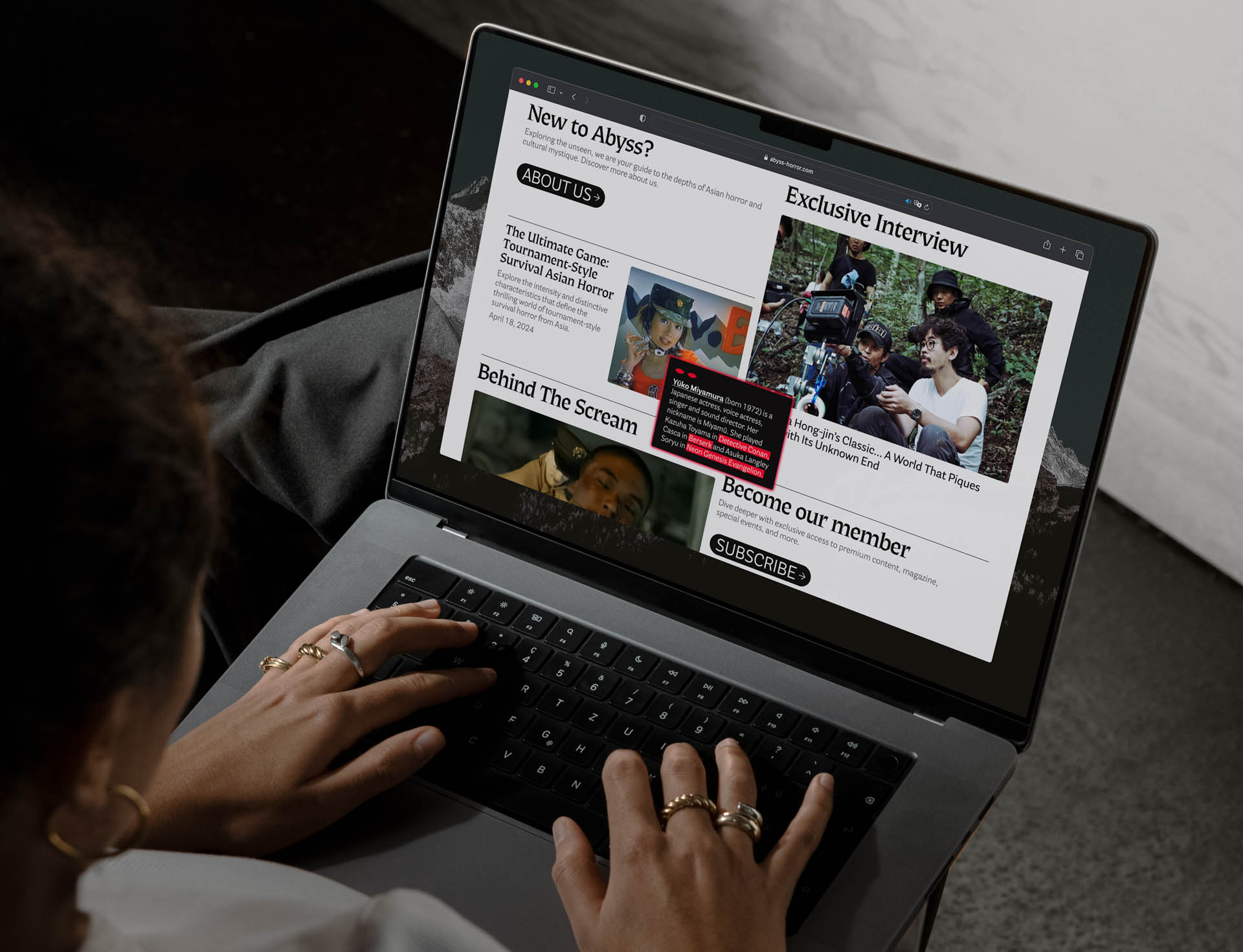 A person with dark hair and multiple rings on their fingers is using a silver laptop. The laptop screen displays a webpage titled “Exclusive Interview” featuring images and text related to “Ayass,” including a headline “The Ultimate Game-Changing Survival Horror” and “Behind the Scram.” Below the interview text, the page includes a “Subscribe” button. The person’s hands are positioned on the laptop’s keyboard as they interact with the screen.
