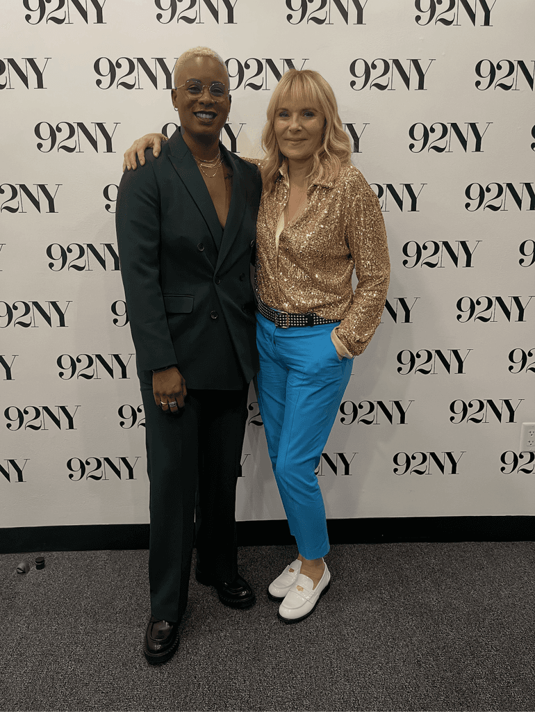 Ashleigh Warren and Kim Cattrall pose together in front of a branded backdrop, smiling and dressed in stylish outfits.