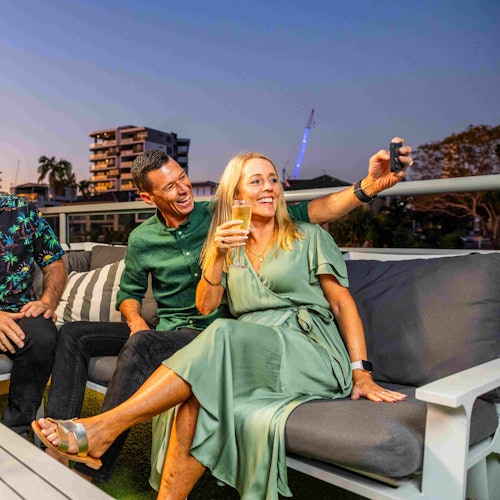 Three people sitting on a rooftop patio; a woman in a green dress takes a selfie with a man next to her. Urban buildings in the background.