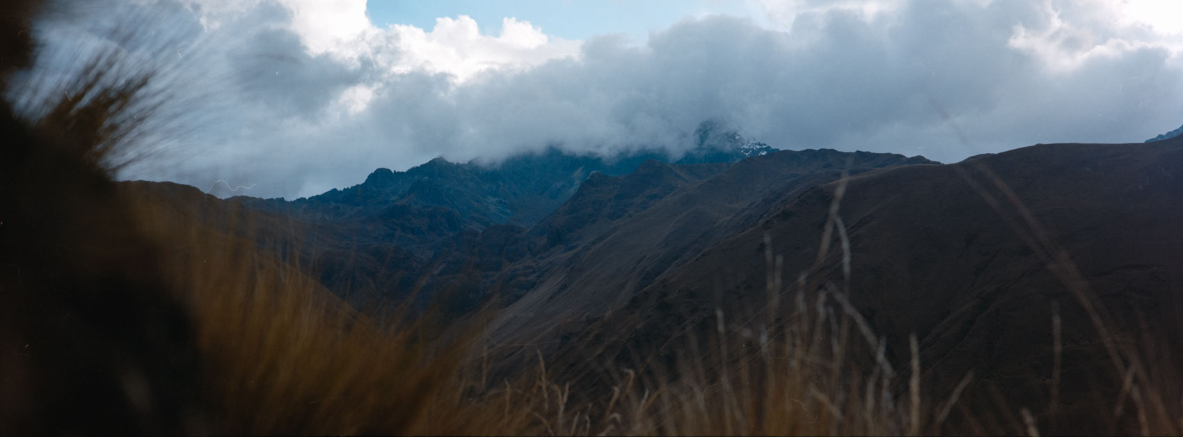 Panoramic film photograph taken near Machu Picchu elevation, showing the mountain peaks covered in dark clouds in the Andean mountains, highlighting everyday life at high altitude.