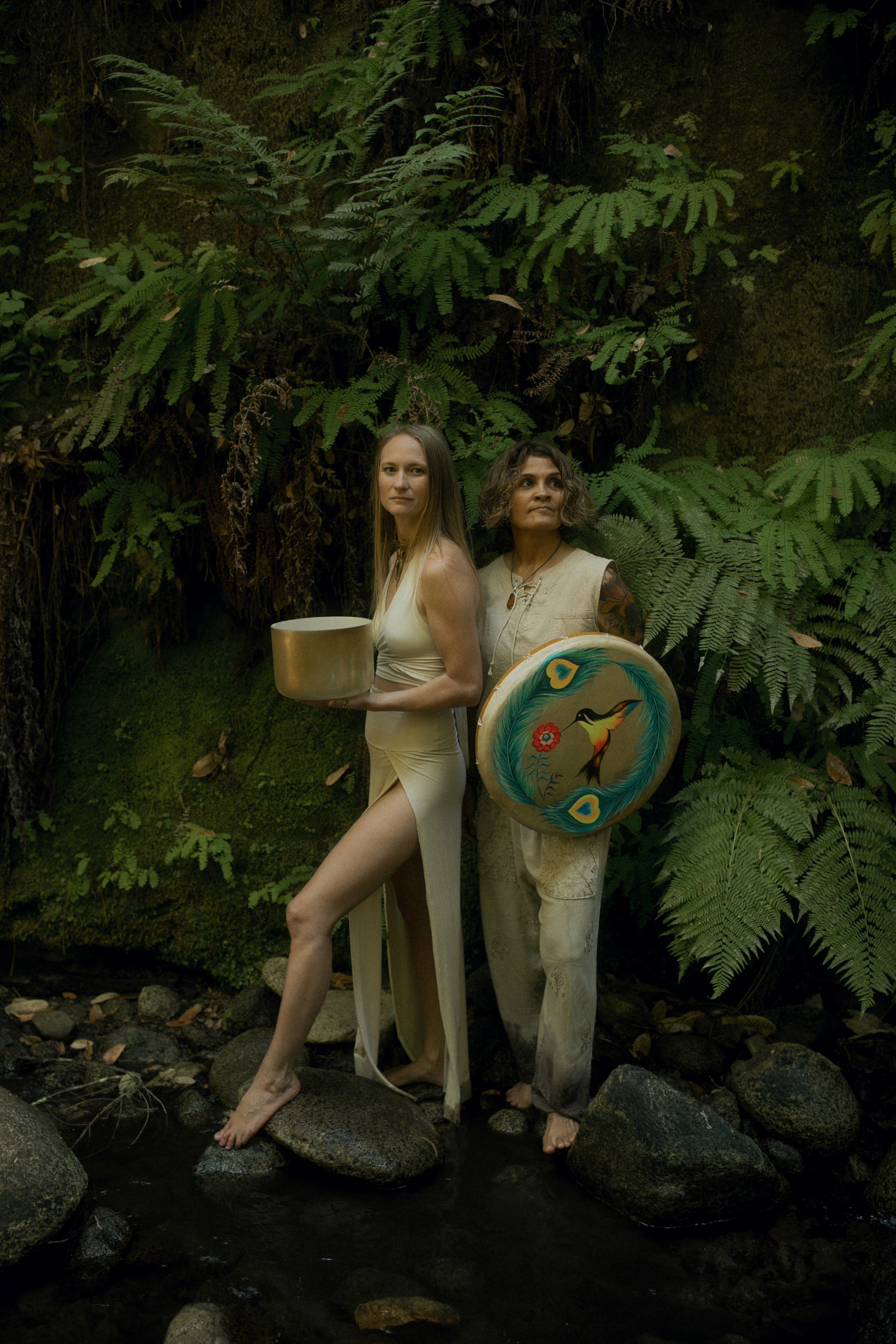 Emeline and Sonia standing back to back in fern-lined jungle holding ceremonial drums for the Bufo Immersion Retreat