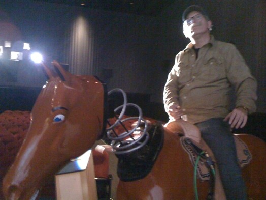 A photo of a man in a theatre, sitting on an artificial brown horse with his head attached by a string to the body.