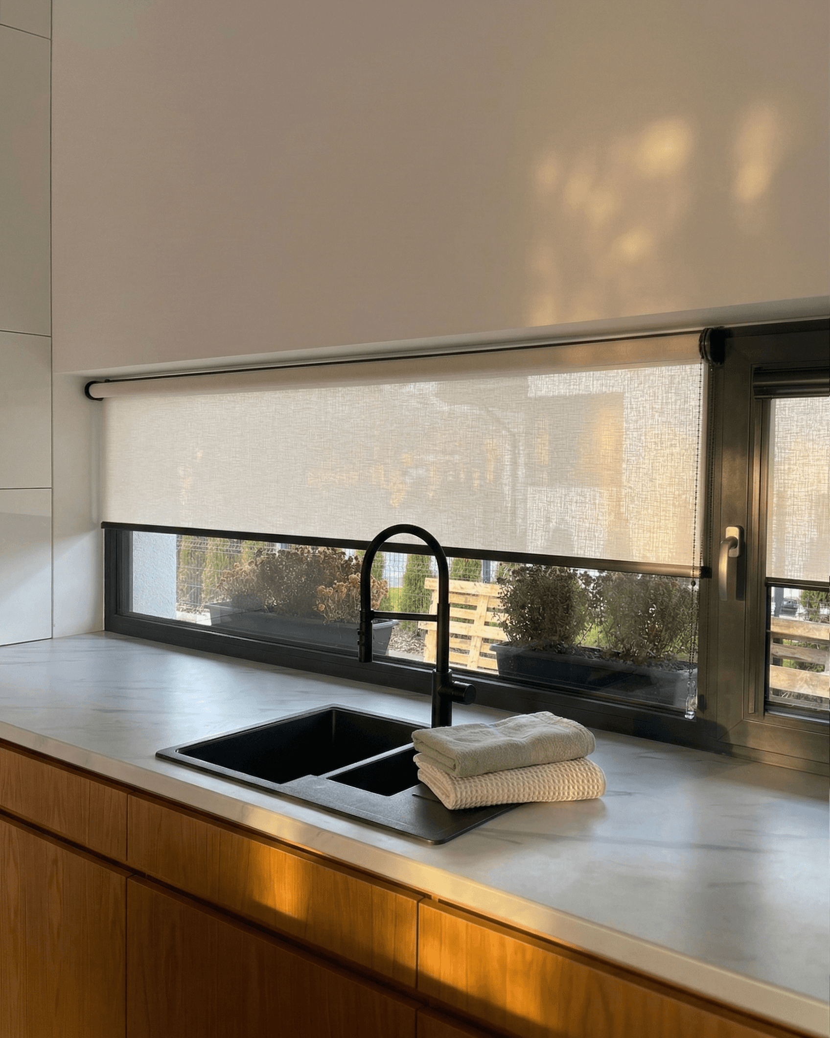 Jasna, półtransparentna roleta materiałowa w nowoczesnej kuchni z niskim, szerokim oknem nad drewnianym blatem. HeyShade Pruszków – osłony okienne na wymiar idealne do kuchni.