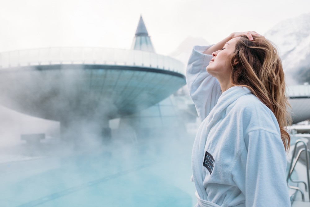 Eine Frau im weißen Bademantel steht neben einem dampfenden Außenpool im Aqua Dome Therme Längenfeld, umgeben von schneebedeckten Bergen und nebligem Himmel.