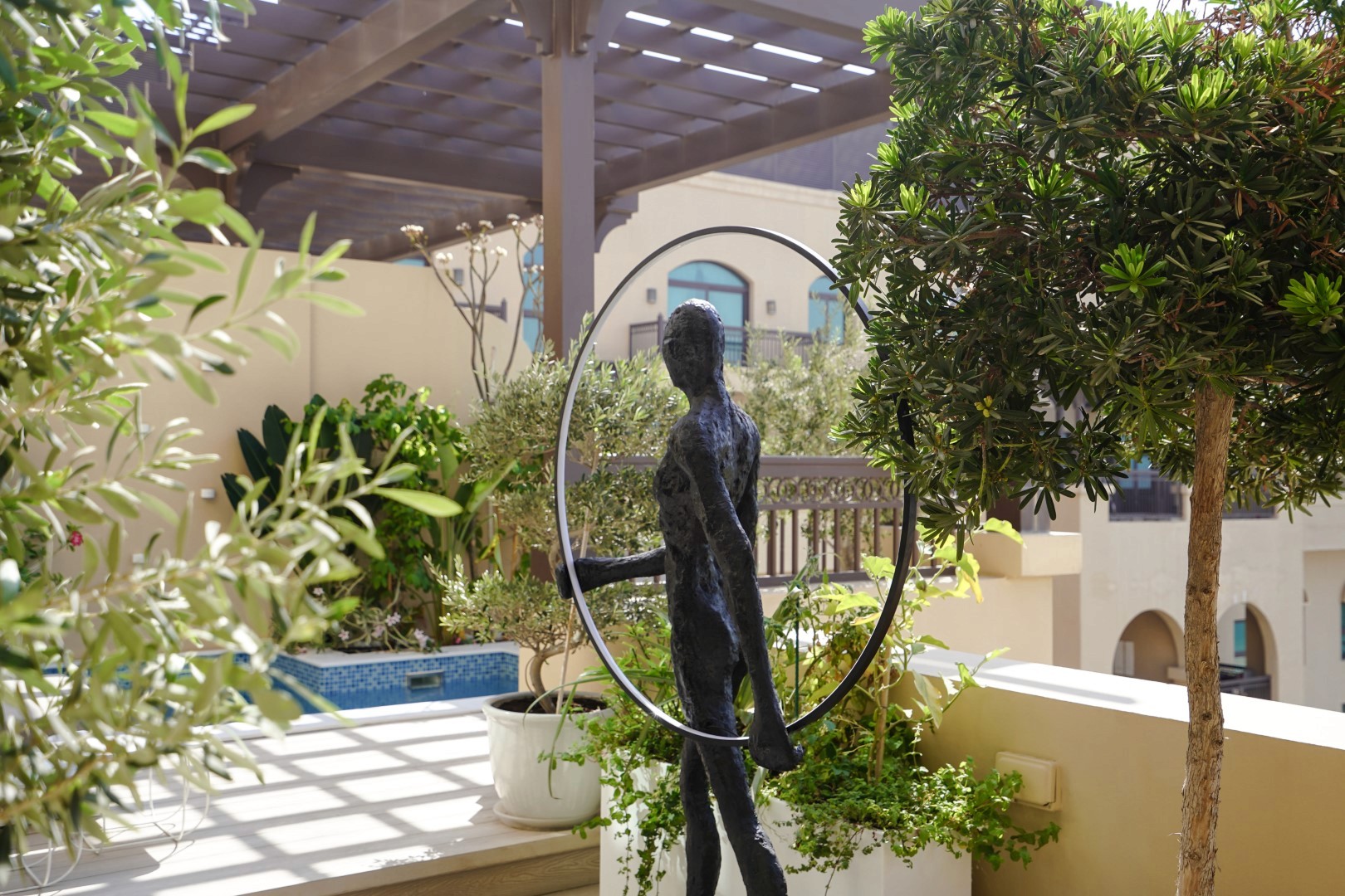A contemporary sculpture of a human figure is set amidst lush greenery and positioned on a sunlit patio in District 1, Dubai, surrounded by Mediterranean-style architecture and a pergola, creating a serene outdoor art display.