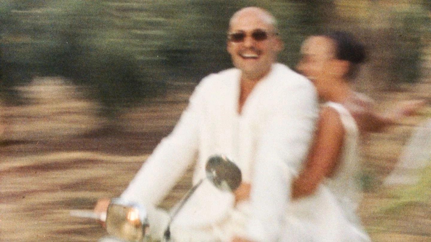A man and woman dressed in white are joyfully riding a scooter through a blurred, sunny outdoor setting, capturing a sense of motion and happiness.