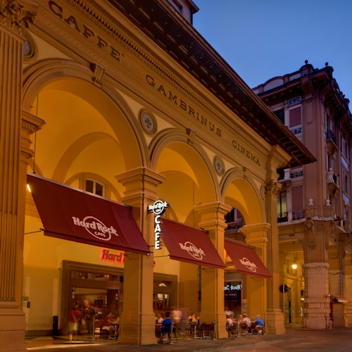 An illuminated café with arches and "Caffè Gambrinus Cinema" signage. People seated under red "Hard Rock Café" awnings.