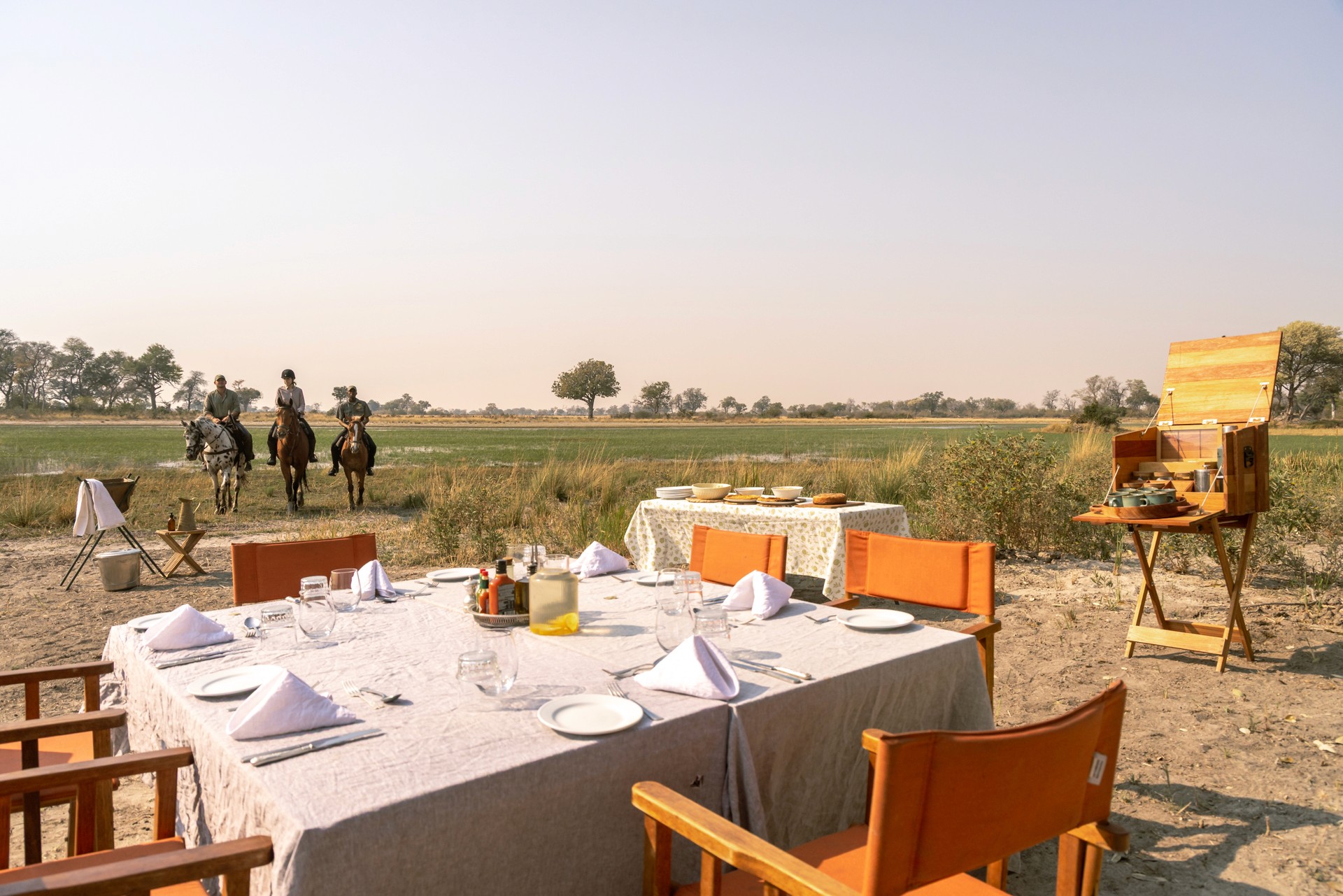 Kujwana Delta Ride, Okavango Delta, Botswana. Tre ryttare till häst på ridresa skrittar fram emot ett uppdukat bord mitt i Okavangos vildmark.
