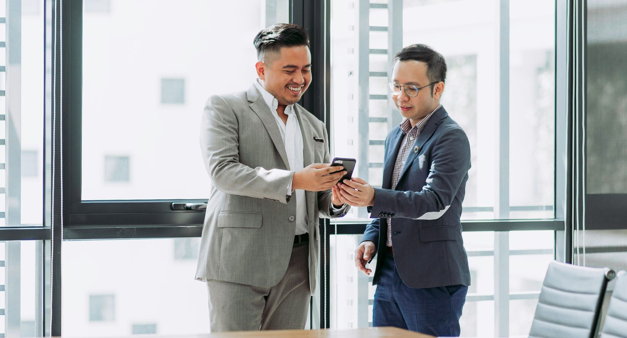 dois homens de terno em um escritório olhando algo aparentemente interessante no celular