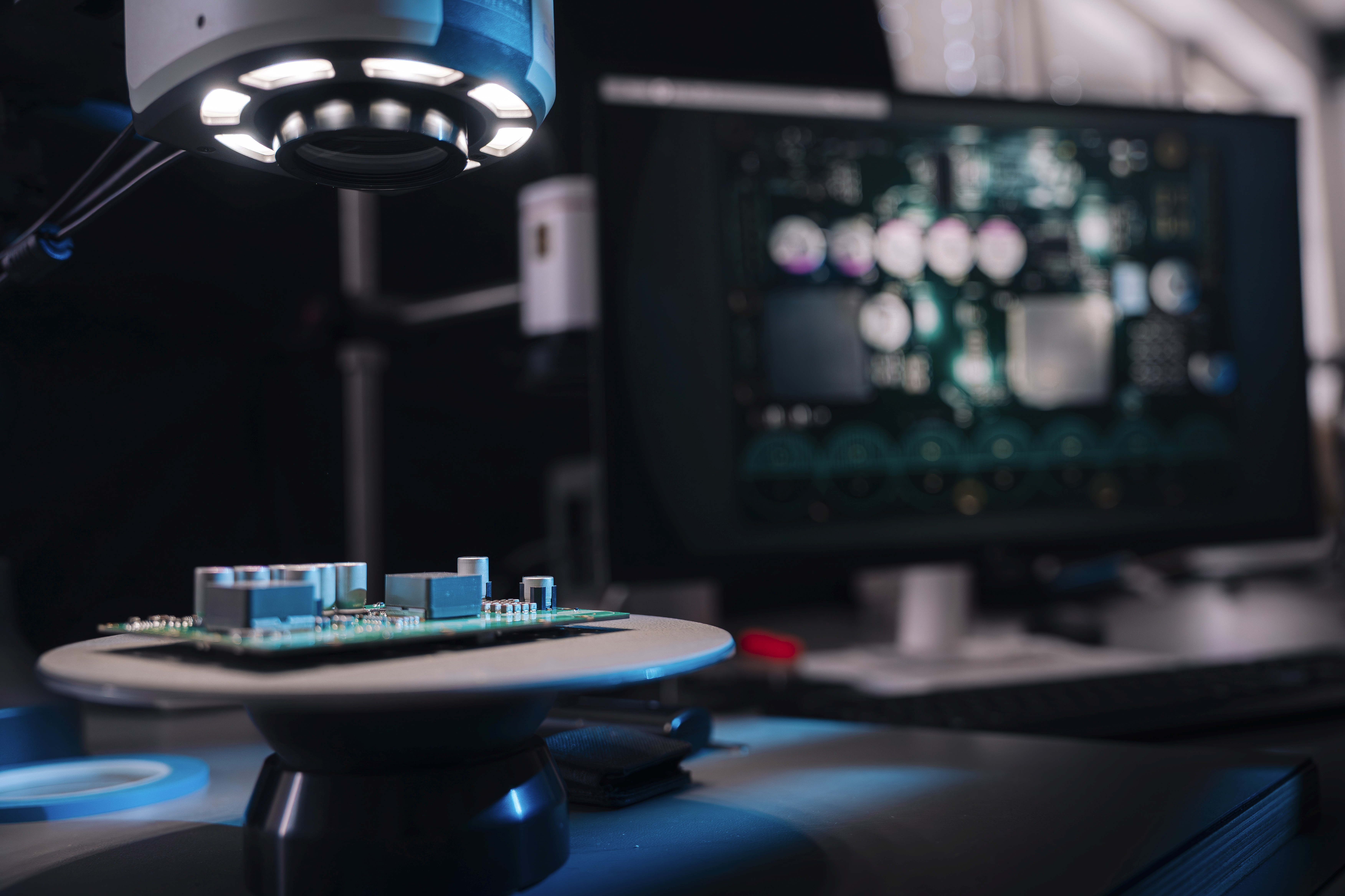 A circuit board on a raised platform, illuminated by a top light on a desk, with blurry screens in the dark background.