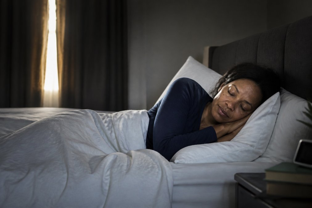 Woman sleeping peacefully in a dark, cool bedroom with soft morning light filtering through blackout curtains, showing proper sleep environment