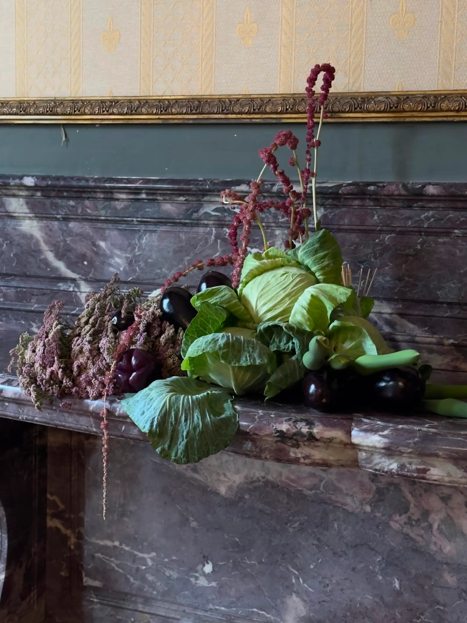 Arreglo decorativo sobre una repisa de mármol con repollos, berenjenas, pimientos y ramas de amaranto rojo, enmarcado por una pared con papel tapiz dorado y molduras clásicas