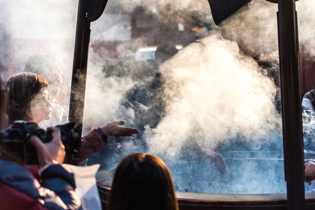 A crowd of people gathered outdoors in a hazy, sunlit atmosphere with thick white smoke drifting across the scene.