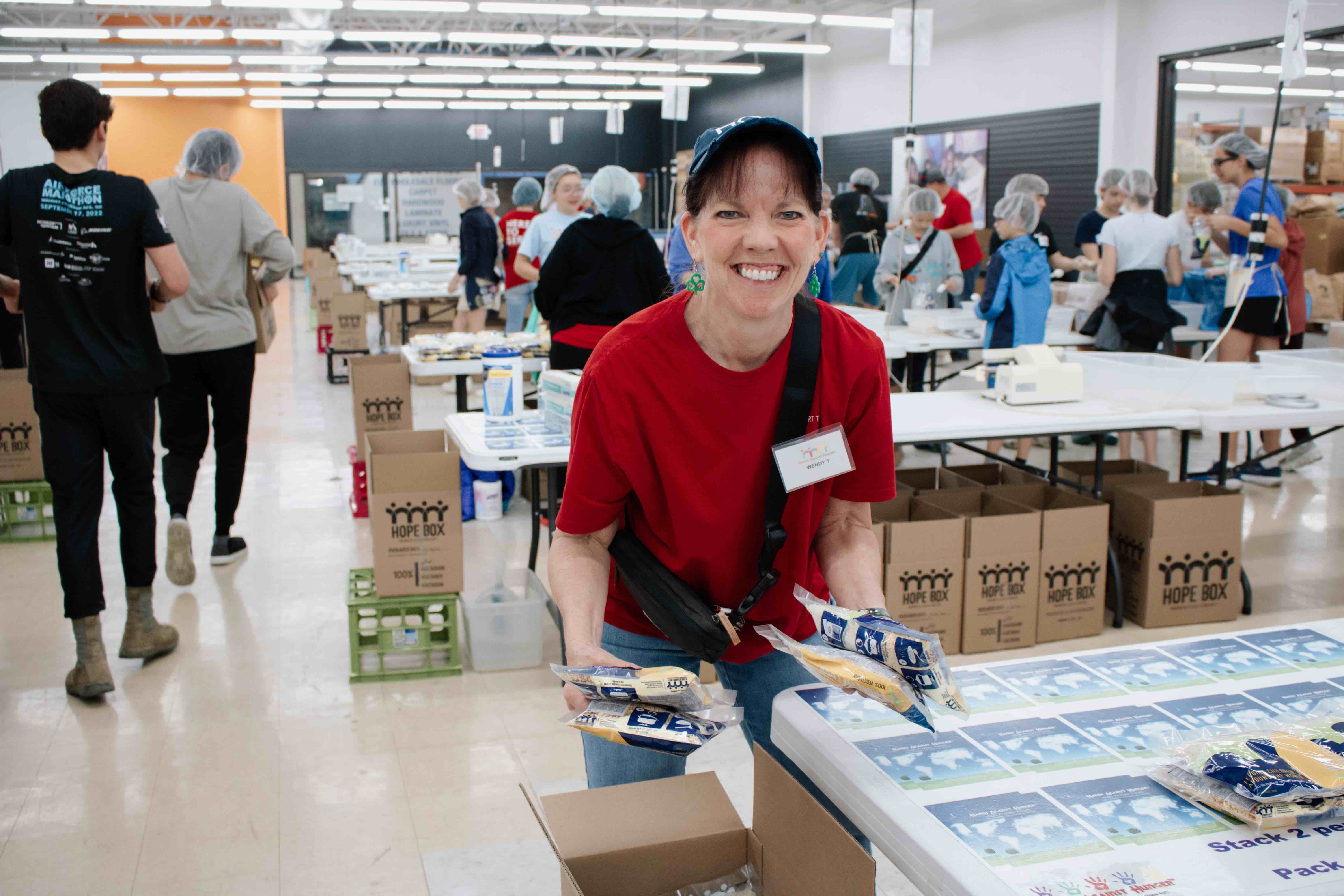 A smiling woman in a red shirt and blue cap packages meal pouches at a Hands Against Hunger® event, surrounded by volunteers and HOPE BOXES.