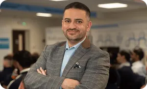 Professional photo of Saj Hussain wearing a grey suit jacket, smiling at camera in an office setting