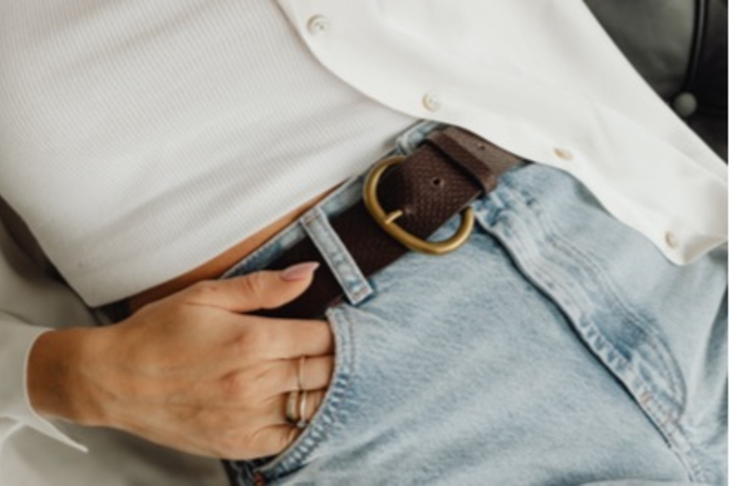 Close-up of a woman wearing light blue jeans, a brown belt with a gold buckle, a white ribbed top and open shirt — showcasing effortless, polished casual styling details.
