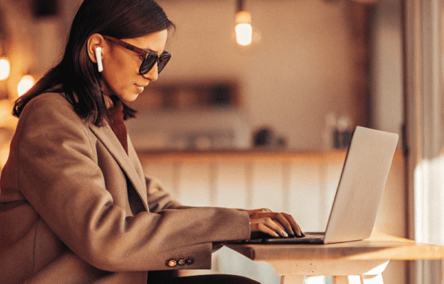 Woman in sunglasses working on a laptop at a cafe. She wears earbuds and a blazer.
