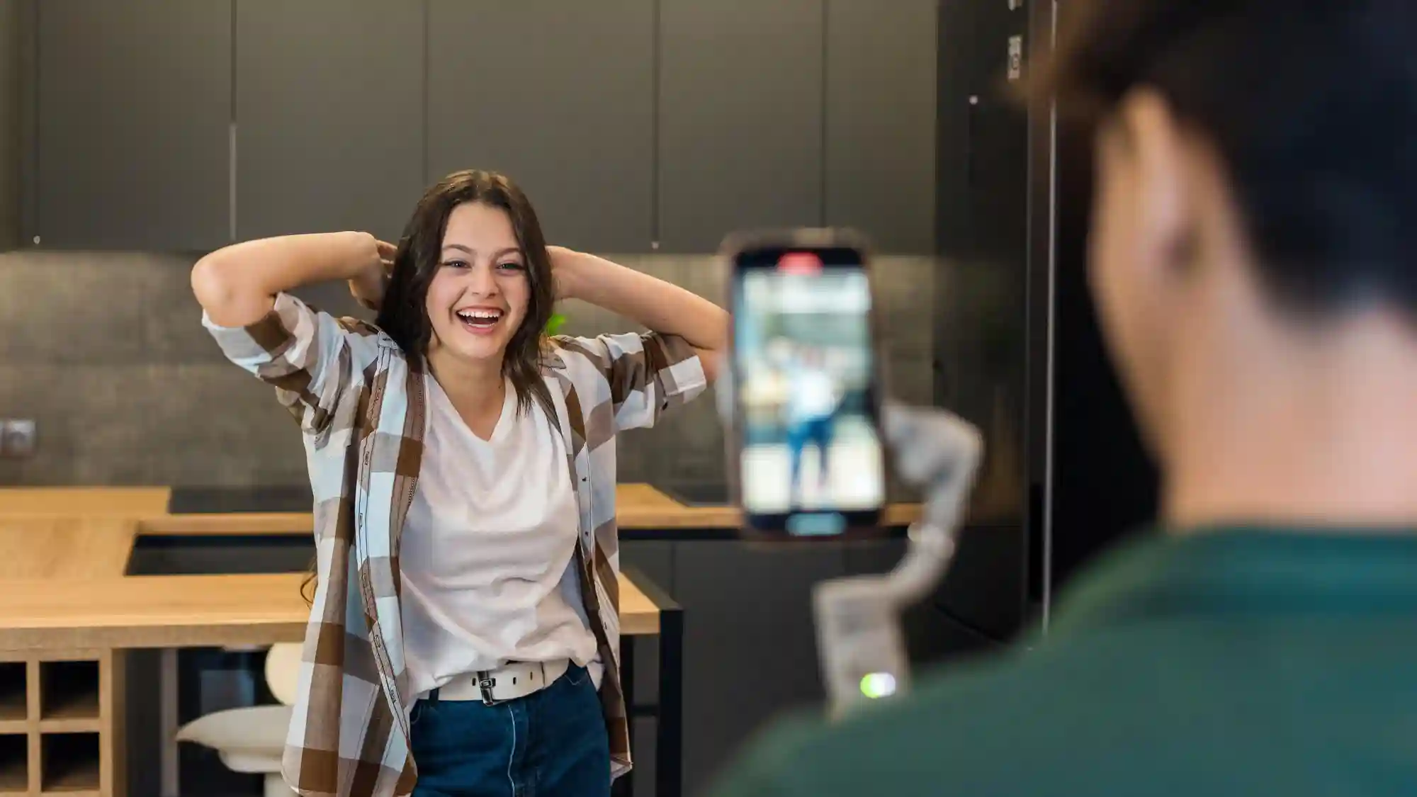 A young woman smiles and poses in a kitchen while a friend records her using a smartphone and gimbal.