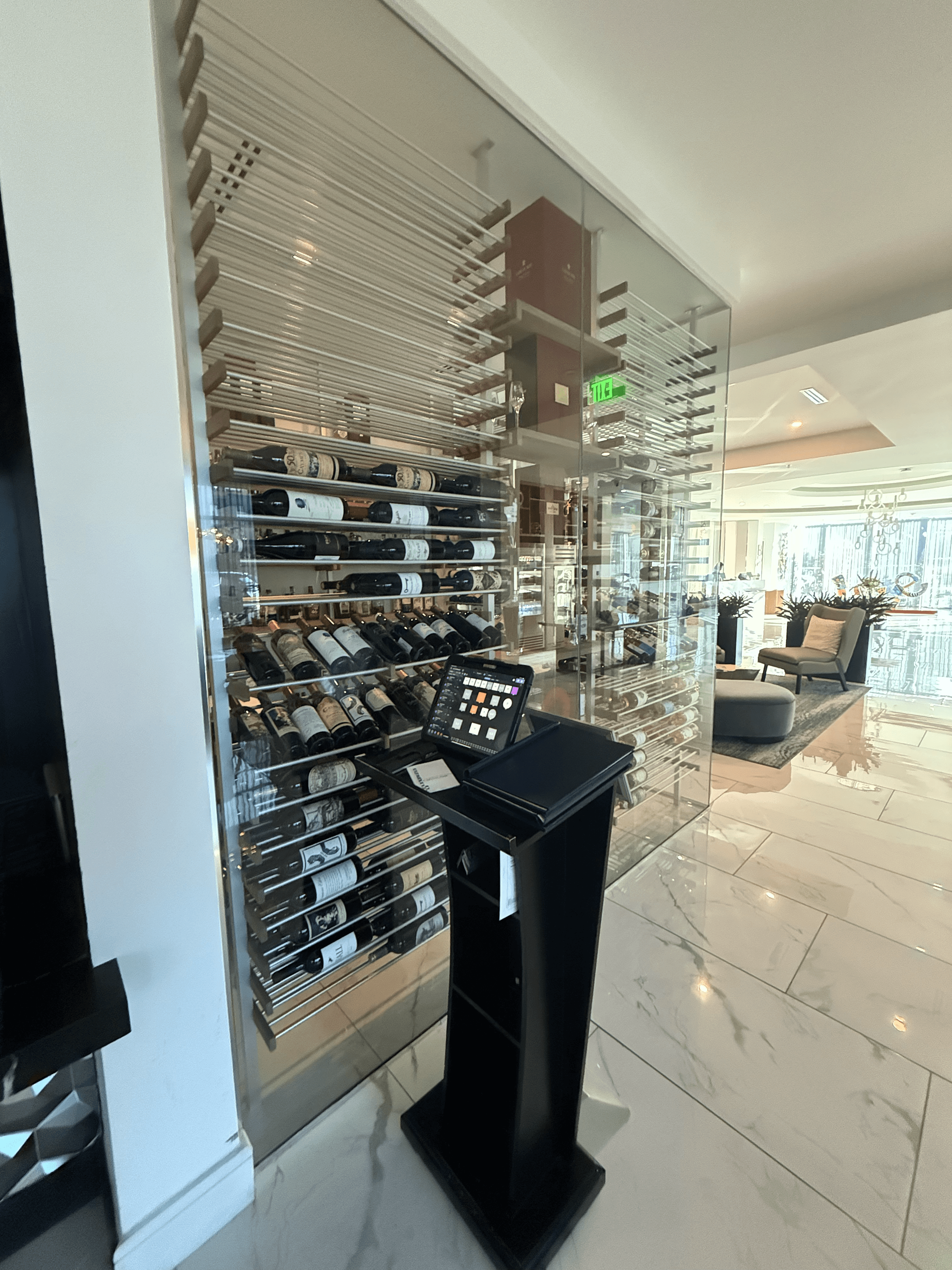 Exterior view of the Cambria Waco Hotel glass-enclosed wine cellar with frameless glass walls, brushed aluminum racking, and walnut detailing beside the restaurant host stand.