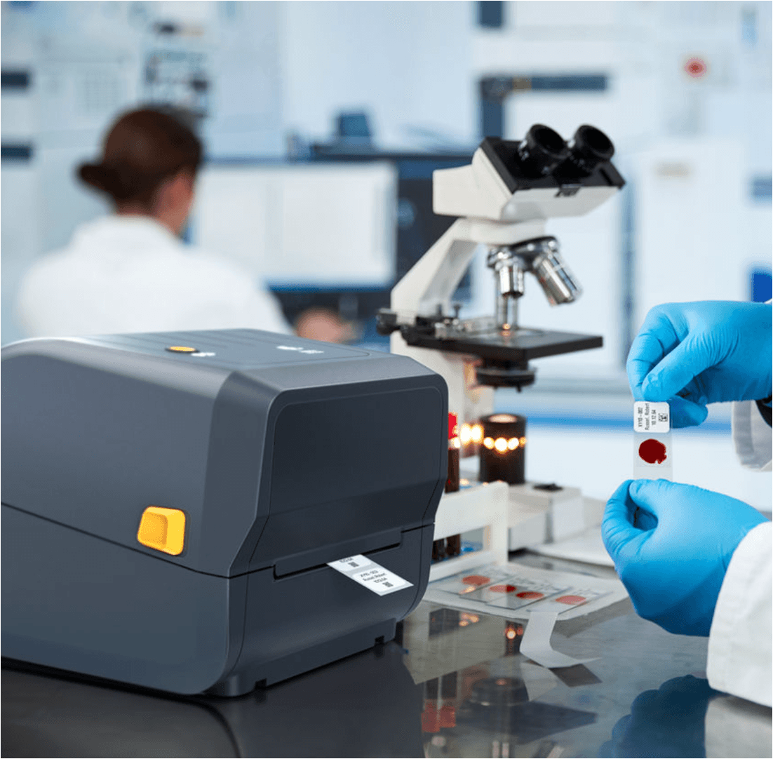 A compact thermal printer printing a QR code label on a desk near plants.